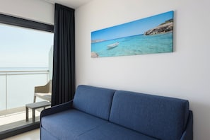 Een moderne kamer met een blauwe bank, een schilderij van een kustlandschap en uitzicht op de zee door een raam.