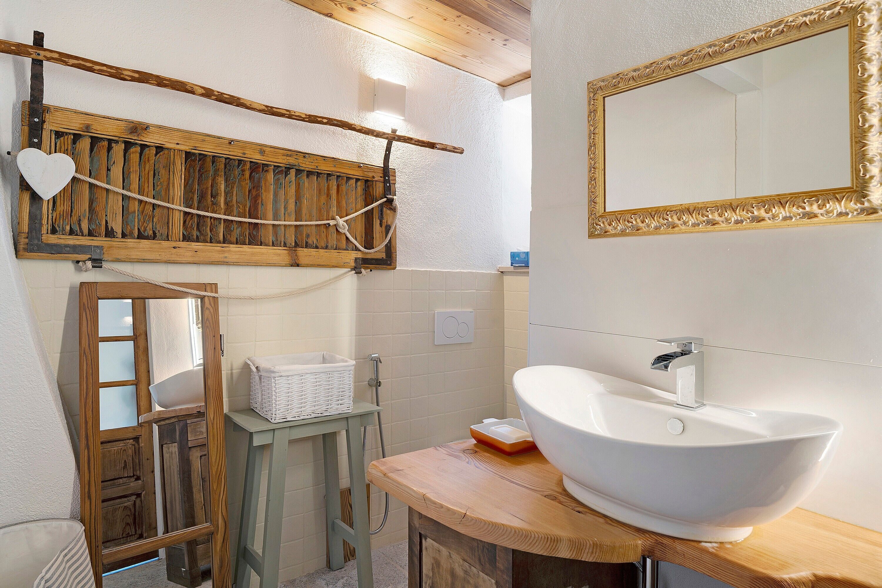 Une salle de bain avec un meuble-lavabo en bois, un lavabo blanc, un miroir avec un cadre doré et un porte-serviettes en bois orné d’un motif en forme de cœur.