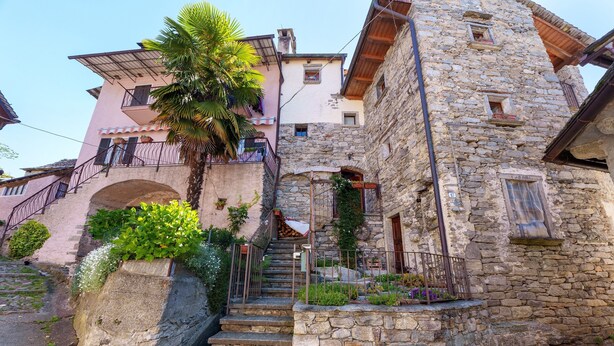 Ein Gebäude aus Stein mit einer Treppe, die zu einem Balkon führt, umgeben von Grünpflanzen und einem rosafarbenen Gebäude im Hintergrund.