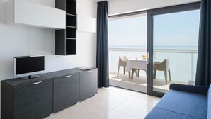 A modern room with a flat-screen TV on a dark cabinet, a balcony with a table and chairs, and a view of the sea. - Beachside flat equipped with everything you need (Lignano Sabbiadoro)