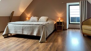 Quarto com teto inclinado, cama com manta texturizada, duas luminárias de cabeceira, mesa de cabeceira de madeira, uma cadeira e uma janela com cortinas.
