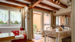 Ein gemütliches Zimmer mit Holzbalkendecke, einem Esstisch mit Tischdecke, einer Couch mit roten Kissen und einem Fenster mit Blick auf einen Garten.