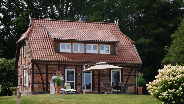 A two-story house with a red-tiled roof, a brick exterior, and a covered patio with outdoor furniture. - Unique half-timbered house on a park-like plot with fantastic views (Südheide)