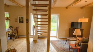 Un escalier en bois menant à un deuxième étage, un coin repas avec des chaises et une table, un téléviseur posé sur un meuble, et un canapé avec une lampe.