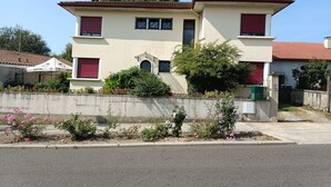 A two-story building with a red-tiled roof, a small front garden with pink flowers, and a concrete wall in front. - The Trumpets of Jericho (Tartas)