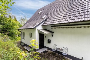 Una casa con techo inclinado, un pequeño porche con mesa y sillas, y un jardín con plantas.
