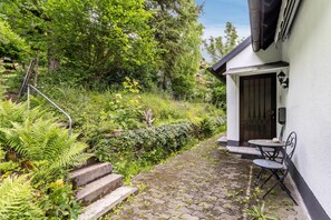A stone pathway leading to a white building with a dark door, a small outdoor table and chairs, and steps ascending to the entrance.