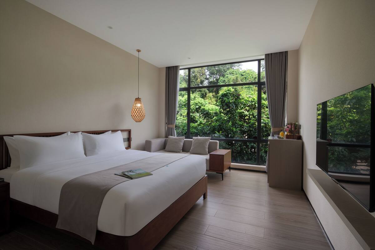 Habitación de hotel moderna con una cama grande, televisión, zona de estar y vistas a la vegetación a través de amplios ventanales.