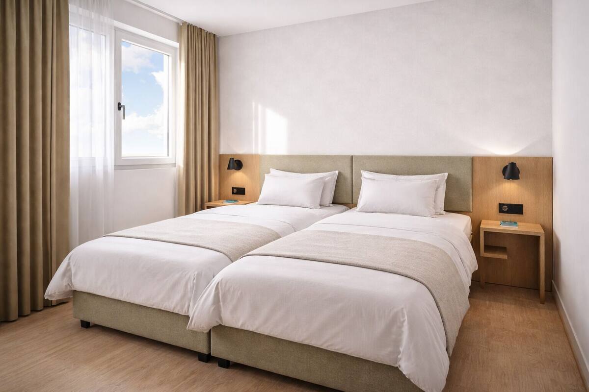 Uma cama de casal com duas mesas de cabeceira, uma janela com cortinas e uma parede lisa.