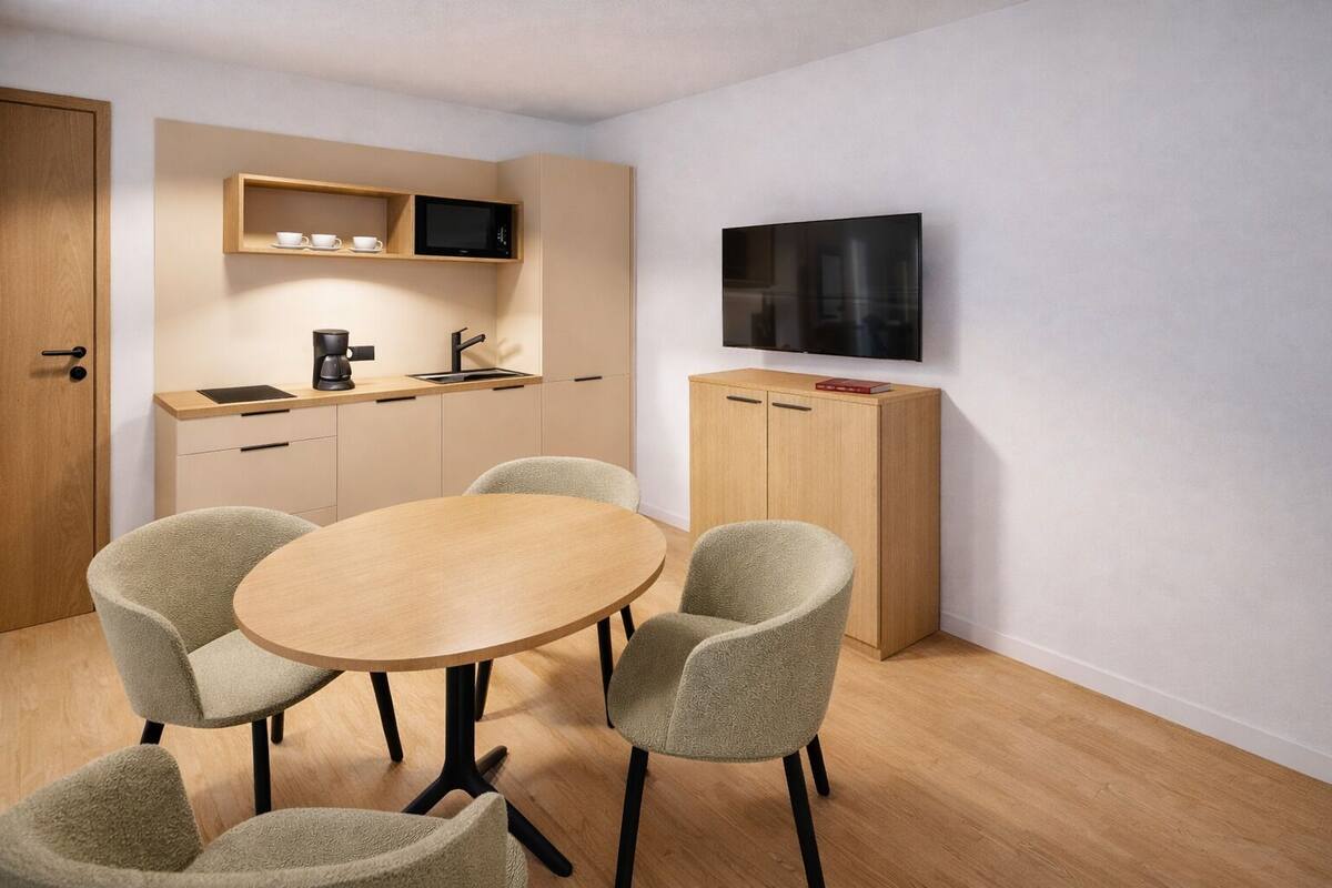 Um espaço compacto com cozinha compacta, área de refeições com mesa redonda e quatro cadeiras, e televisão fixada na parede.