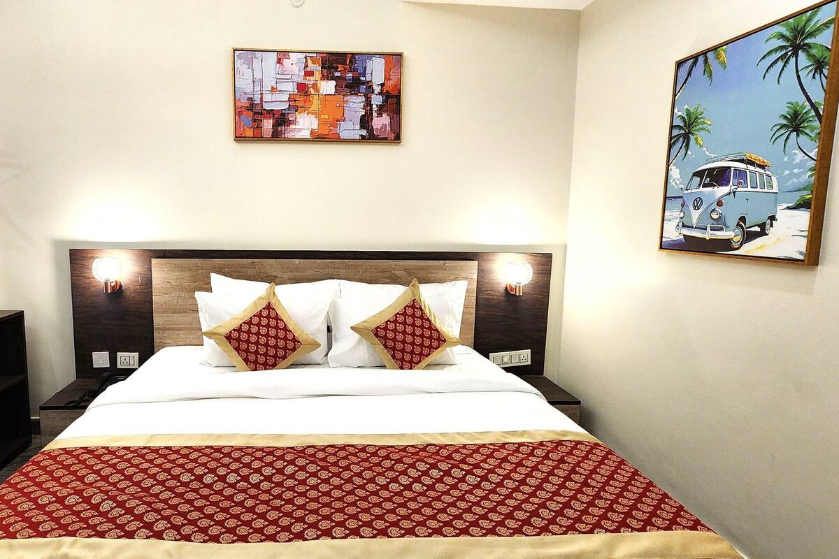 A hotel room with a bed, wooden headboard, two pillows, a red patterned bedspread, a wall-mounted abstract painting, and a framed beach scene painting.