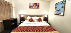 A hotel room with a bed, wooden headboard, two pillows, a red patterned bedspread, a wall-mounted abstract painting, and a framed beach scene painting. - RoadRolls (Varanasi)