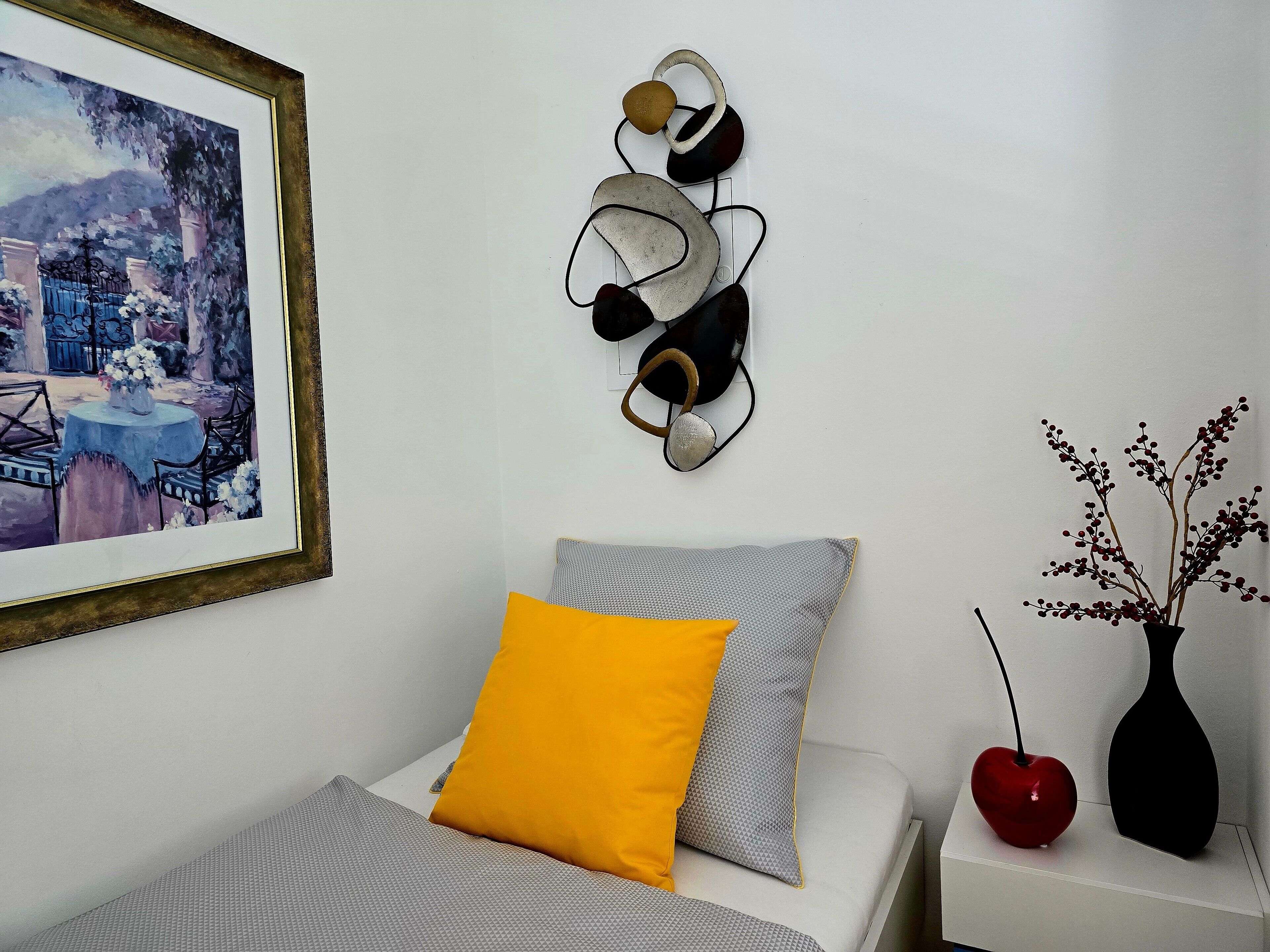 Un lit avec un oreiller gris et jaune, un luminaire fixé au mur et un tableau encadré.