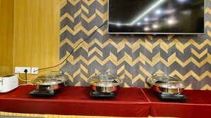 A room with a TV mounted on the wall, a red table with three covered food stations, and a wooden panel on the left. - VN Alpha Suites (Bengaluru)