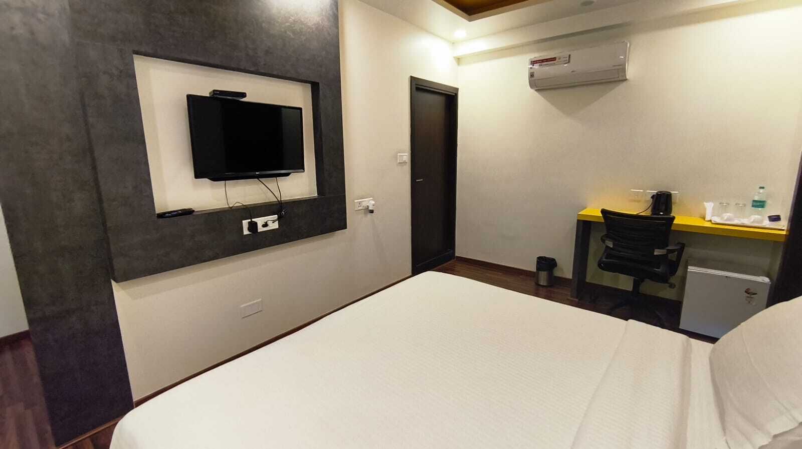 A hotel room with a bed, a desk with a chair, a television mounted on the wall, and an air conditioning unit.
