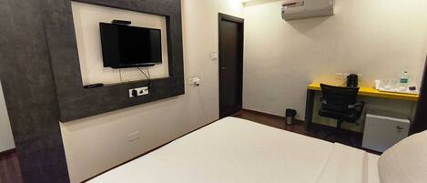 A hotel room with a bed, a desk with a chair, a television mounted on the wall, and an air conditioning unit.