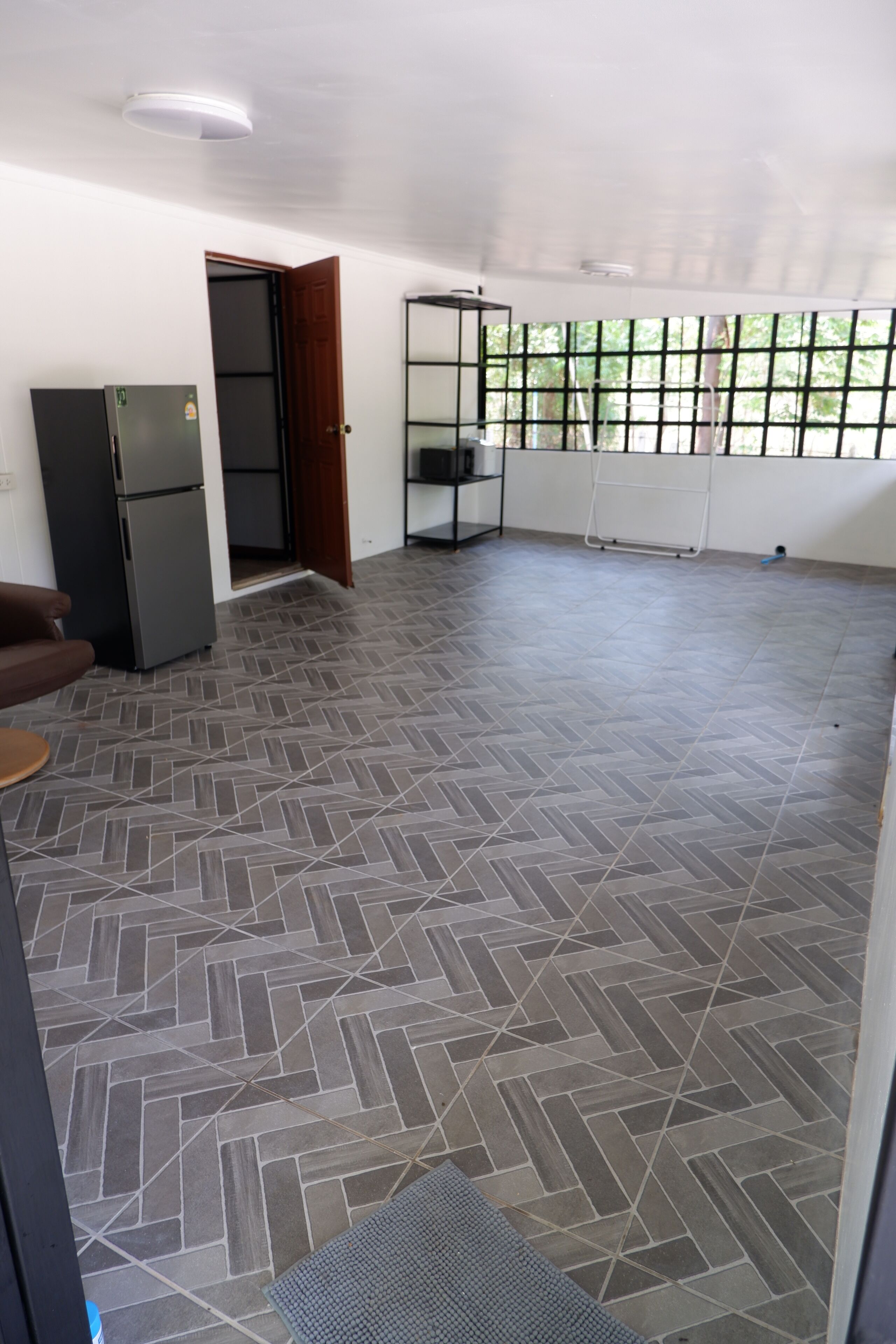 A spacious room with a herringbone-patterned floor, a refrigerator, a door leading to another room, and a window with a view of greenery.