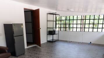 A spacious room with a herringbone-patterned floor, a refrigerator, a door leading to another room, and a window with a view of greenery.