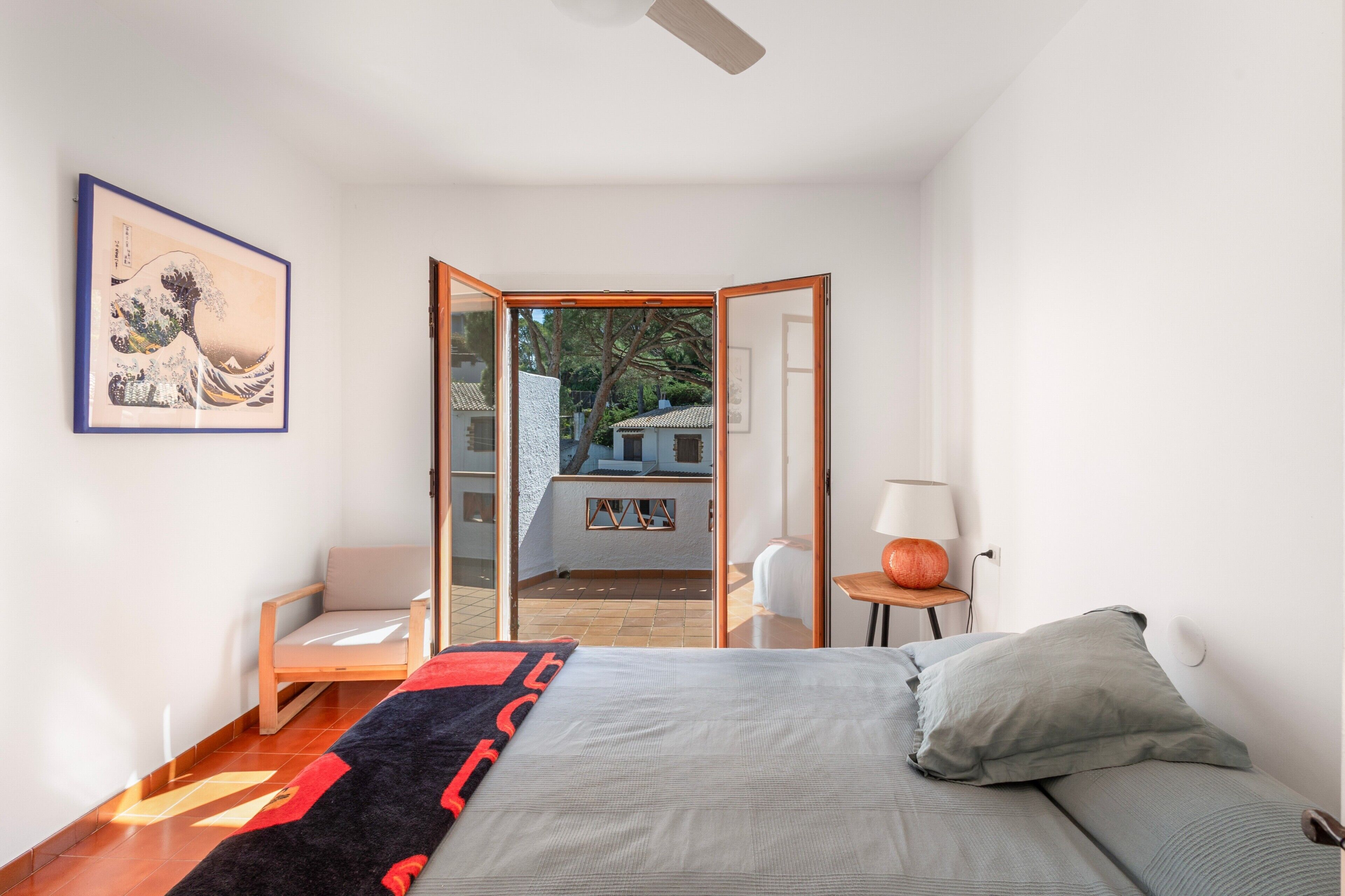 Een slaapkamer met een bed, een stoel, een tafel, een lamp en een lijst met een afbeelding aan de muur.