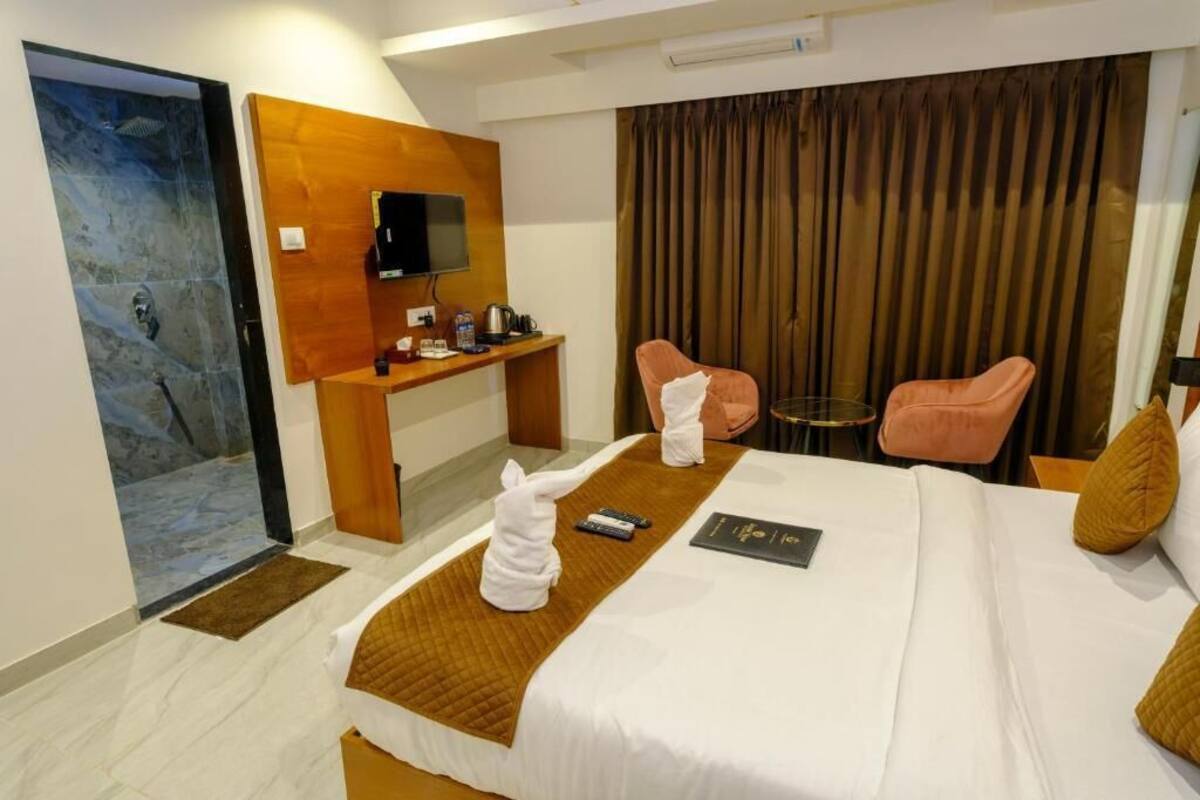Quarto de hotel com cama, escrivaninha, televisão, duas cadeiras e chuveiro.