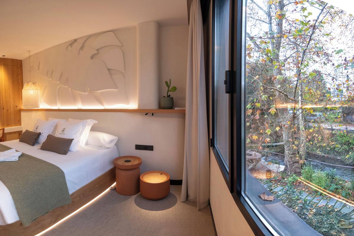A modern hotel room with a large bed, bedside tables, a desk, a wall-mounted light, and a view of trees through a window.