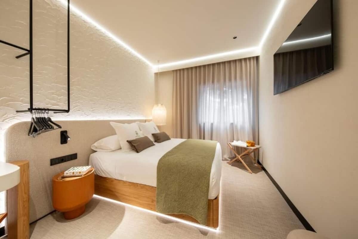 A modern hotel room with a neatly made bed, a bedside table with a lamp, a small wooden stool, and a television mounted on the wall.