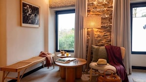A cozy room with a stone wall, a bench, a round table with a book and glasses, and a chair with a hat. - Hotel Sarela Natura (Santiago de Compostela)