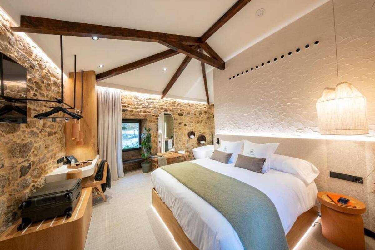 A bedroom with a stone wall, a bed with a green blanket, a wooden desk with a phone, a mirror, and a hanging light fixture.
