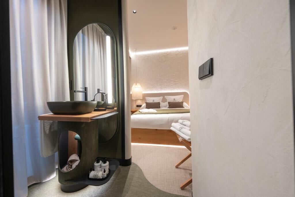 A modern hotel room with a large round mirror, a wooden side table with a bowl, a bed with white linens, and a bedside table with a lamp.