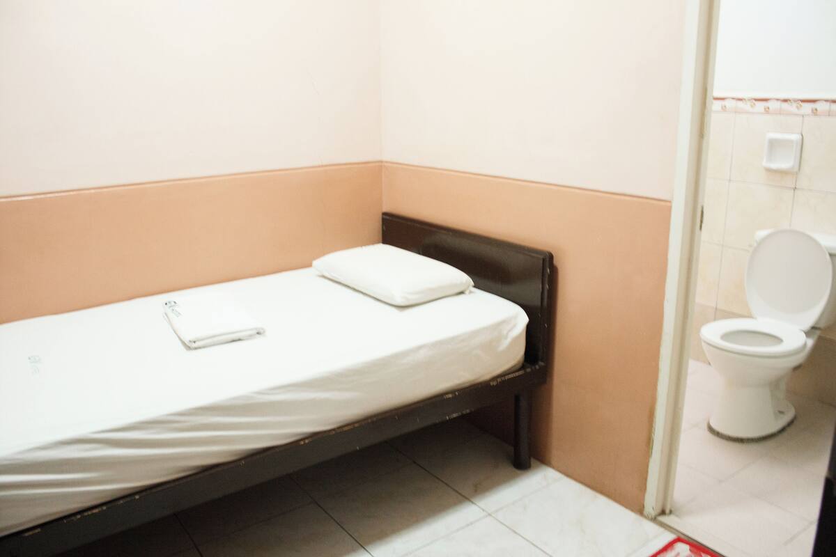 Ein kleines Zimmer mit einem Einzelbett, einer weißen Matratze, einem schwarzen Kopfteil und einem weißen Kopfkissen. Rechts befindet sich eine weiße Toilette und eine kleine, weiße Ablage am Wand.