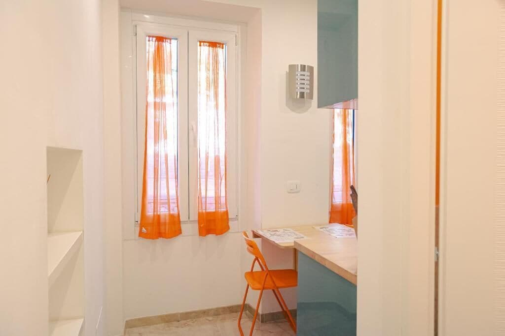 Ein kleiner Raum mit Schreibtisch, Stuhl und einem Fenster mit orangenen Vorhängen.