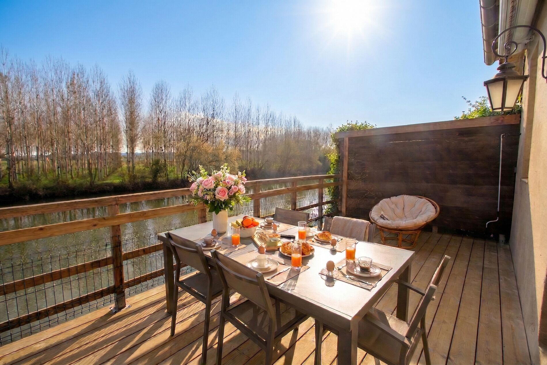 Une terrasse en bois avec une table dressée pour le petit-déjeuner, offrant une vue sur un plan d’eau et des arbres.
