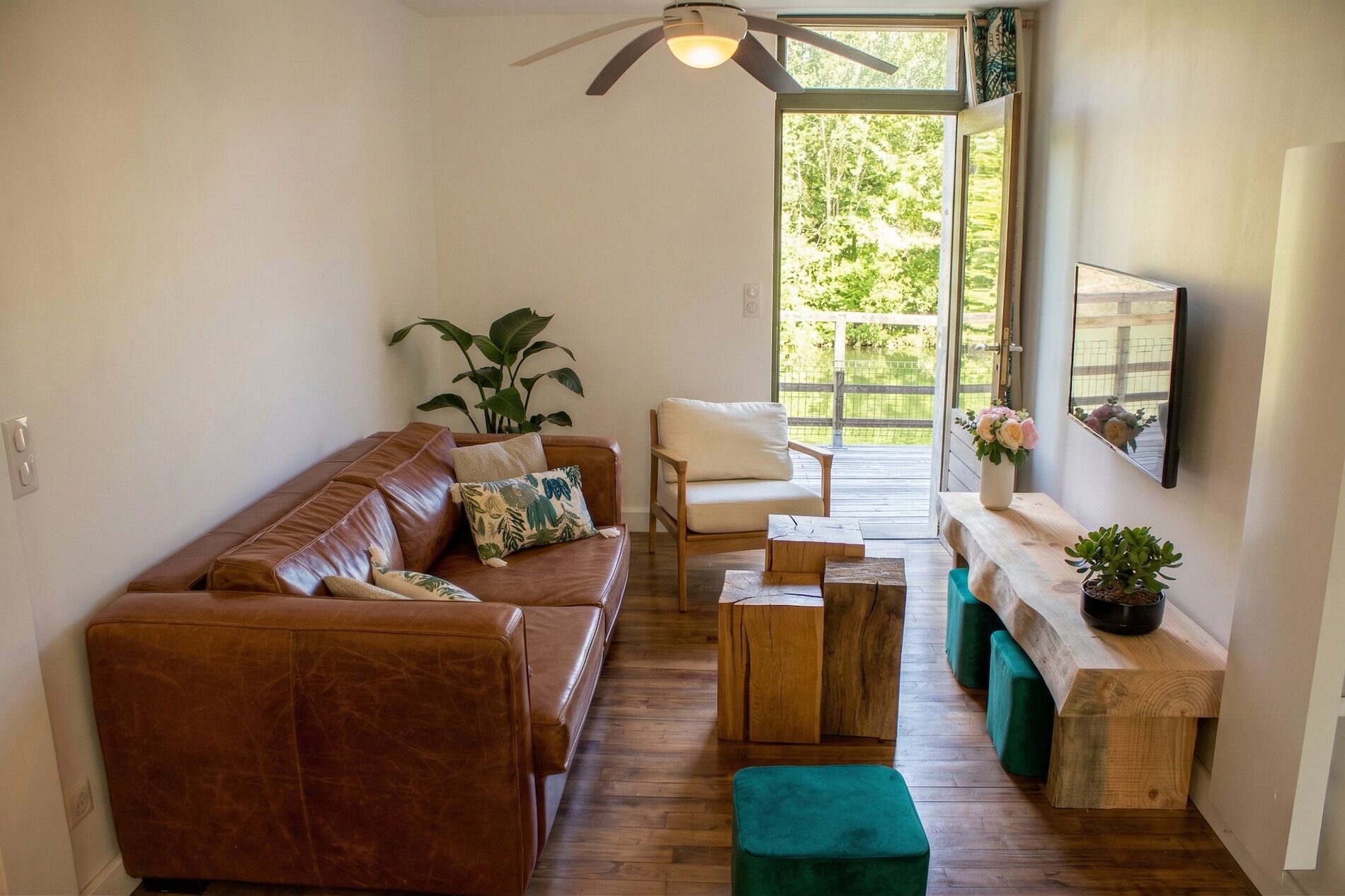 Un salón con un sofá de cuero marrón, una mesa de centro de madera y un otoman verde. Hay un banco de madera con plantas en macetas, una silla blanca y un ventanal con vistas a zonas verdes.