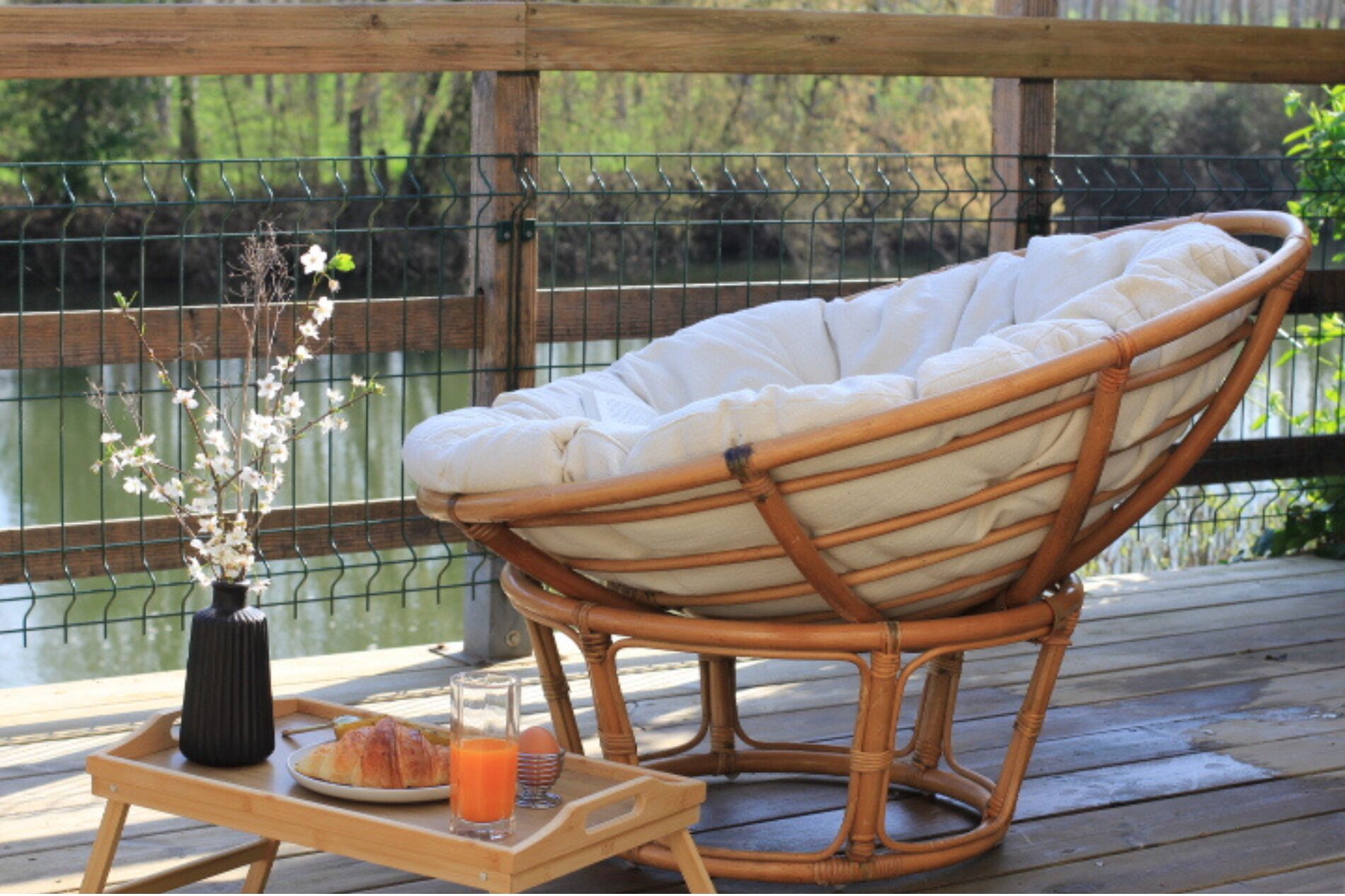 Une chaise en osier avec un coussin, posée sur une terrasse en bois, une petite table avec une assiette de nourriture et un verre de jus d’orange, et un vase rempli de fleurs.