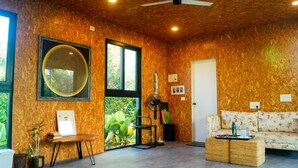 A room with wooden walls, a large window, a fan, a sofa, and a table with a plant. - Amram (Shoolagiri)