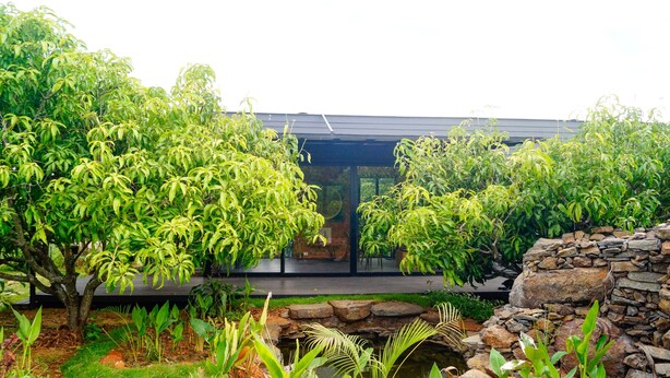 A modern building with a glass facade, surrounded by lush greenery and a stone wall. - Amram (Shoolagiri)
