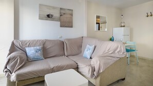Ein beiges Sofa mit zwei Zierkissen, ein weißer Beistelltisch, ein kleiner weißer Schrank und ein blauer Sessel in einem neutral gestalteten Raum mit Bildern an den Wänden.