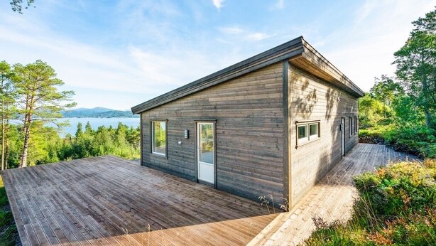 A modern wooden cabin with a deck, large windows, and a view of a lake and mountains.