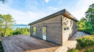 A modern wooden cabin with a deck, large windows, and a view of a lake and mountains.