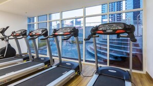 Een sportschool met loopbanden en een hometrainer, grote ramen met uitzicht op gebouwen.