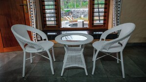 A room with a round table and two wicker chairs, a wooden door, and a window with a view of an outdoor area. - Bodhi (Shoolagiri)