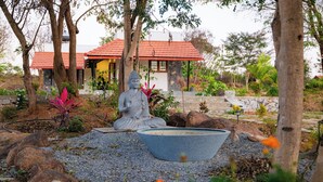 A garden with a Buddha statue, a stone basin, and a house with a red roof. - Bodhi (Shoolagiri)