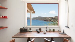 Una habitación con una mesa de madera, un ventanal con vista al mar y una estantería con diversos objetos.