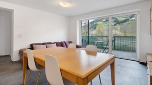 A modern living room with a wooden dining table, white chairs, a sofa with cushions, and a large sliding glass door leading to an outdoor area. - Apartment 'Talferblick Burg' with Mountain View, Private Terrace and Wi-Fi (Sarentino)