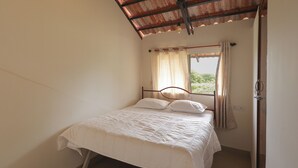 A single bed with a wooden frame and white bedding in a room with a sloped ceiling and a window with curtains. - Arokiya (Shoolagiri)