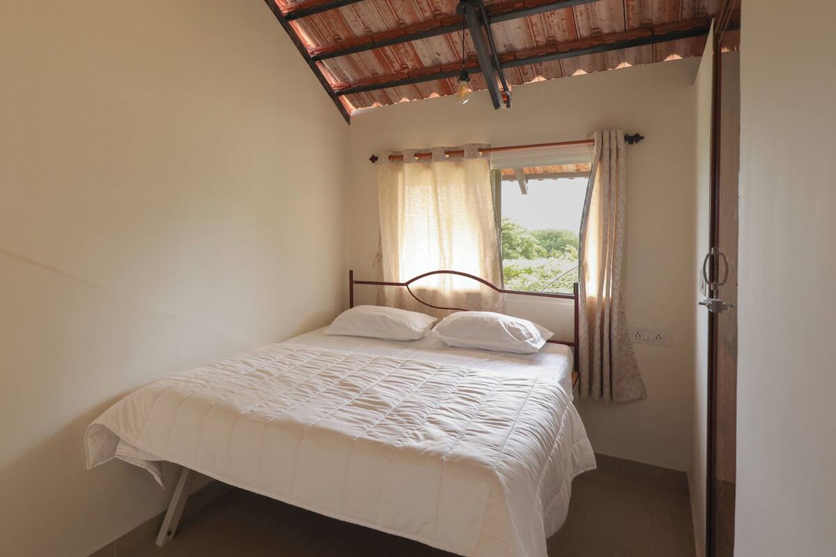 Una cama individual con estructura de madera y ropa de cama blanca en una habitación con techo inclinado y ventana con cortinas.