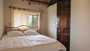 A single bed with a wooden frame and white bedding, a window with curtains, and a door with a decorative panel. - Arokiya (Shoolagiri)