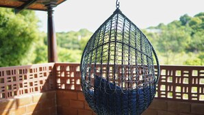 A hanging chair with a netted frame and blue cushion, suspended from a structure with a wooden lattice railing and brick wall. - Laze Daze (Shoolagiri)