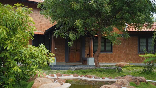 A brick building with a covered porch, a small pond with rocks, and lush greenery. - Laze Daze (Shoolagiri)