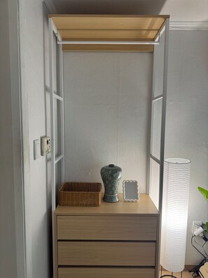 Ein gemütlicher Raum mit einem Holzschrank, einem Korb aus Weidengeflecht, einer dekorativen Vase, einem gerahmten Spiegel und einer hohen weißen Lampe.
